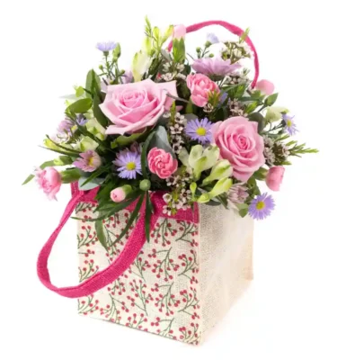 A charming floral arrangement presented in a decorative fabric tote bag with pink handles. The bouquet features soft pink roses, delicate pastel purple daisy-like flowers, petite pink carnations, white freesias, and lush greenery. The tote bag has a cream-colored background adorned with a red berry and green vine pattern, adding a rustic yet elegant touch. This beautifully arranged bouquet is perfect for Mother’s Day, expressing love and appreciation with a fresh and vibrant selection of flowers.
