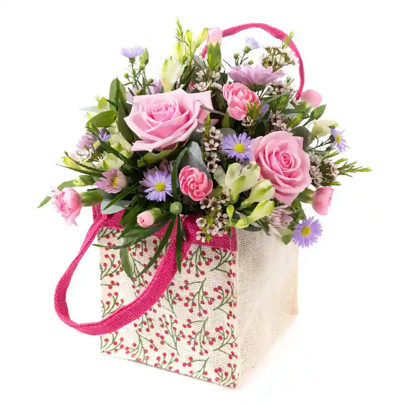 A charming floral arrangement presented in a decorative fabric tote bag with pink handles. The bouquet features soft pink roses, delicate pastel purple daisy-like flowers, petite pink carnations, white freesias, and lush greenery. The tote bag has a cream-colored background adorned with a red berry and green vine pattern, adding a rustic yet elegant touch. This beautifully arranged bouquet is perfect for Mother’s Day, expressing love and appreciation with a fresh and vibrant selection of flowers.
