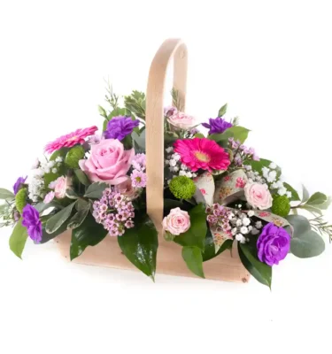 A beautiful floral arrangement in a rustic wooden basket with a curved handle, featuring pink roses, gerbera daisies, purple carnations, green button mums, and delicate white gypsophila, accented with lush greenery and floral-patterned ribbon.
