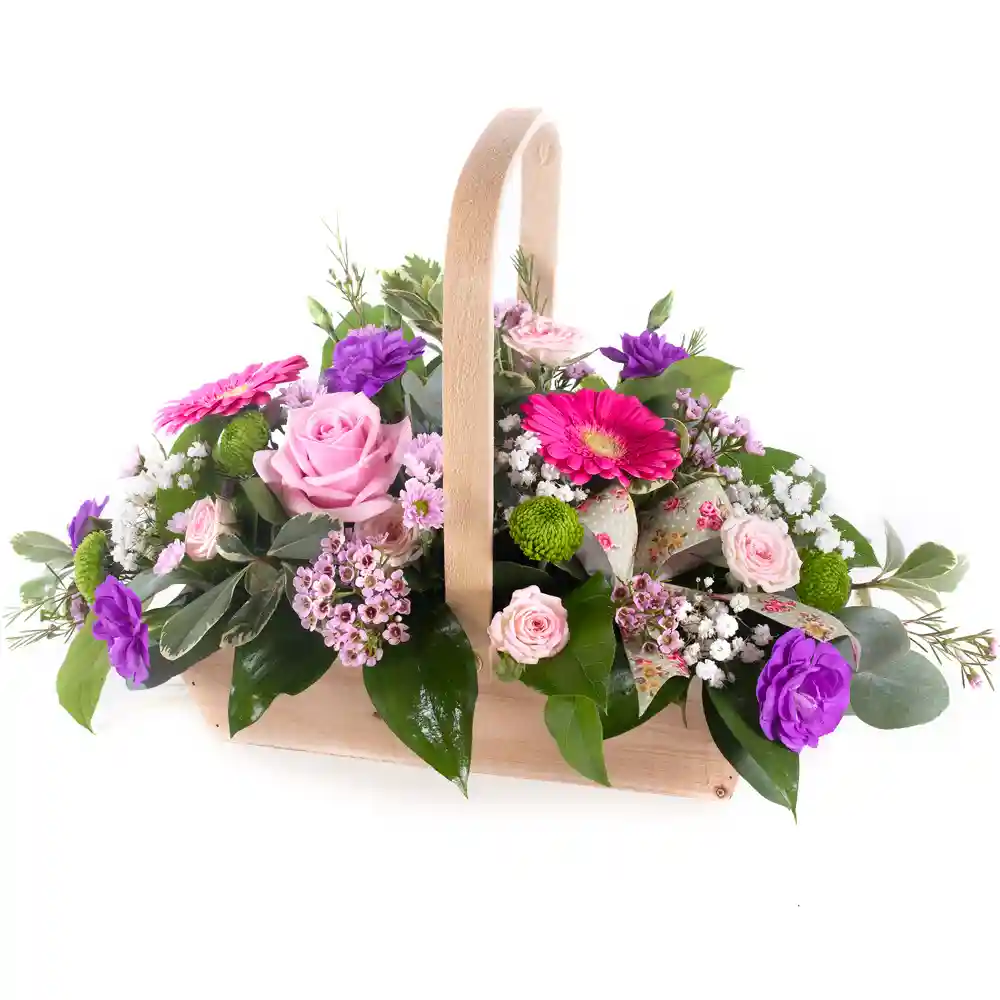 A beautiful floral arrangement in a rustic wooden basket with a curved handle, featuring pink roses, gerbera daisies, purple carnations, green button mums, and delicate white gypsophila, accented with lush greenery and floral-patterned ribbon.