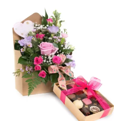 A beautiful Mother's Day bouquet featuring soft pink roses, delicate purple lisianthus, tiny white Gypsophila, and lush green foliage, all arranged in a charming kraft paper envelope. The bouquet is accented with a decorative ribbon and a heart-shaped embellishment. Accompanying the flowers is a box of gourmet chocolates wrapped in a pink ribbon, creating a thoughtful and elegant gift