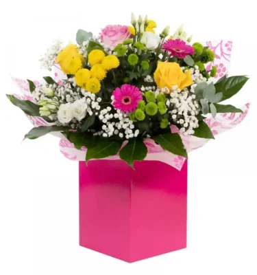A vibrant mixed flower bouquet in a bright pink gift box, featuring yellow roses, pink gerberas, white lisianthus, green chrysanthemums, and delicate gypsophila.