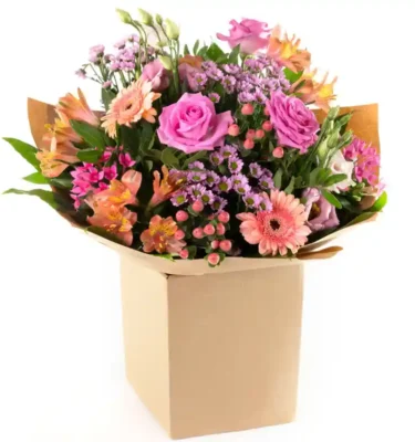 An elegant bouquet of pink, peach, and purple flowers, including roses, gerbera daisies, and wildflowers, arranged with lush green foliage in eco-friendly packaging inside a cardboard box.