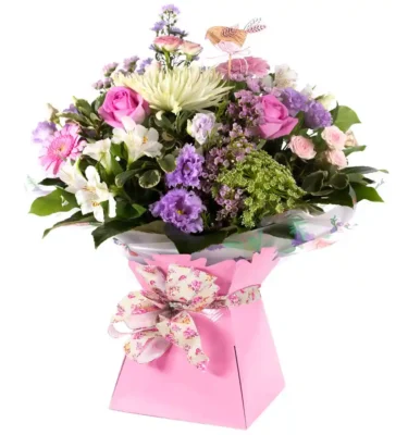A delicate pastel floral bouquet arranged in a soft pink gift box, featuring pink roses, lilac lisianthus, white chrysanthemums, pink gerbera daisies, and sprigs of greenery. The bouquet is wrapped in clear decorative paper and adorned with a floral-patterned bow, with a small wooden bird decoration nestled among the blooms.
