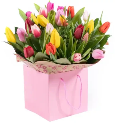 A vibrant bouquet of multicolored tulips, including shades of yellow, pink, red, and orange, elegantly arranged with lush green leaves in a soft pink gift bag with floral-patterned wrapping.