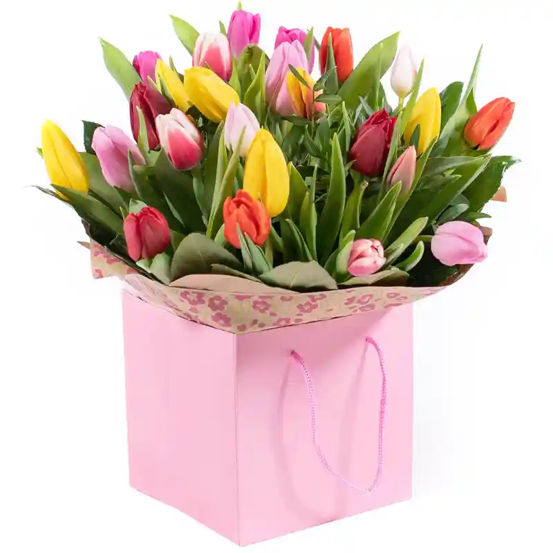 A vibrant bouquet of multicolored tulips, including shades of yellow, pink, red, and orange, elegantly arranged with lush green leaves in a soft pink gift bag with floral-patterned wrapping.